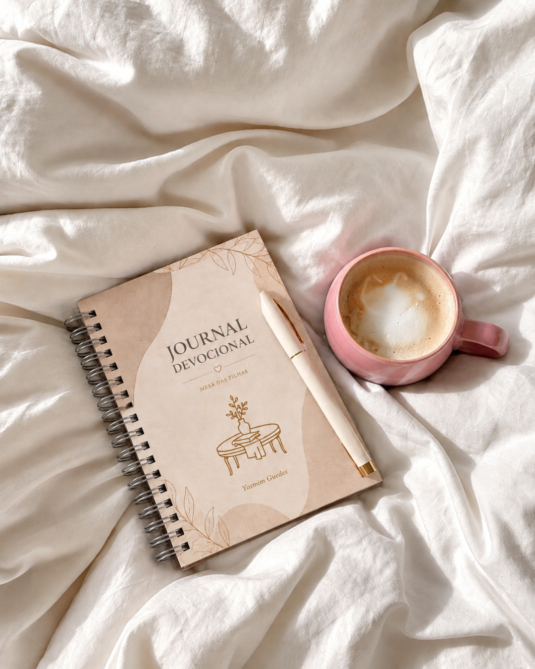 Journal devocional em tecido rosé com detalhes dourados sobre superfície creme